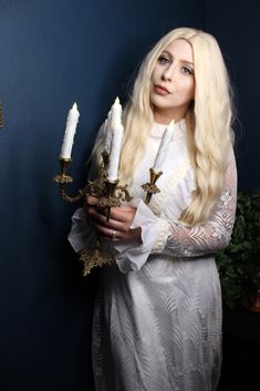 a woman with long blonde hair holding two candles in her hands and wearing a white dress