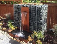 a water fountain in the middle of a garden with rocks and plants around it, next to a wooden fence