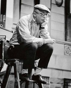 a man sitting on top of a wooden stool in front of a building with his hand under his chin