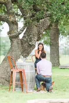 a man and woman sitting at a table under a tree next to a wine barrel