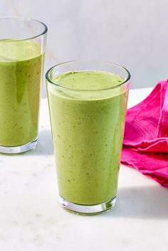 two glasses filled with green smoothie sitting on top of a white counter next to a pink napkin