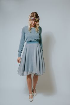 a woman in a blue sweater and grey skirt is standing against a white wall with her hands on her hips