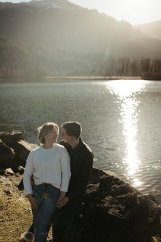 a man and woman standing next to each other near the water with mountains in the background