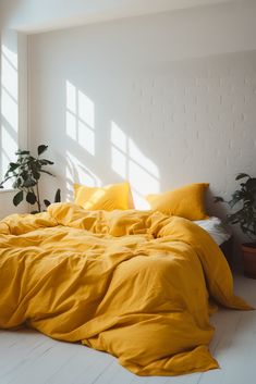 an unmade bed with yellow sheets and pillows in a white room next to potted plants