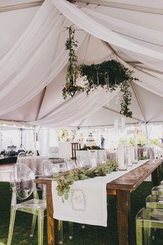 an outdoor tent set up with clear chairs and greenery hanging from the ceiling above