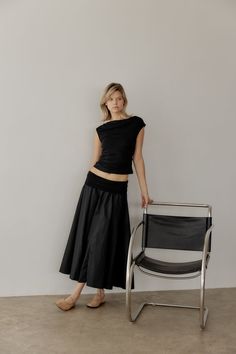 a woman standing next to a chair in front of a white wall, wearing a black top and skirt