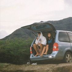 two people sitting in the back of a van on top of a dirt hill with mountains in the background