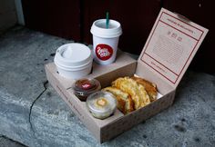 an open box with some food and drinks on the ground next to a cup of coffee