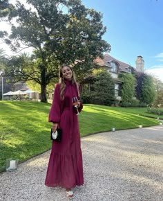 a woman in a red dress is standing on the gravel road outside her house and smiling at the camera