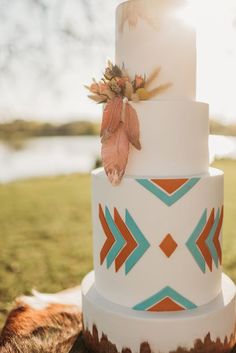 a wedding cake with an orange and blue design on it sitting in the grass next to a lake