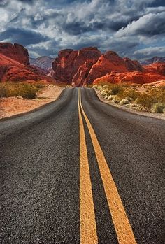 an empty road in the desert with mountains in the backgrouund and clouds in the sky