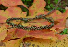 a green and brown braided bracelet sitting on top of leaf covered ground next to leaves