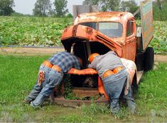 two men working on an old truck in the mud, with weeds growing out of it