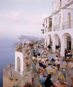 many people are sitting at tables outside by the water and some have drinks in their hands