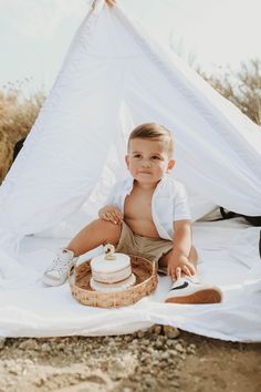 a little boy sitting in front of a white tent with a cake on the ground