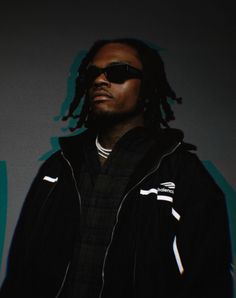 a man with dreadlocks standing in front of a wall wearing sunglasses and a black jacket