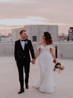 a newly married couple holding hands and walking in an open space with the sun setting behind them