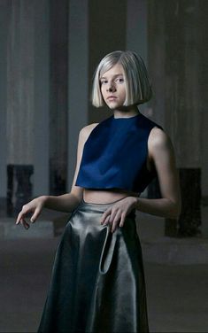 a woman with blonde hair wearing a blue top and black skirt standing in an empty room