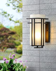 an outdoor wall light on the side of a building with flowers in front of it