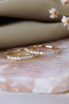 two gold wedding bands with diamonds sitting on top of a pink marble surface next to flowers