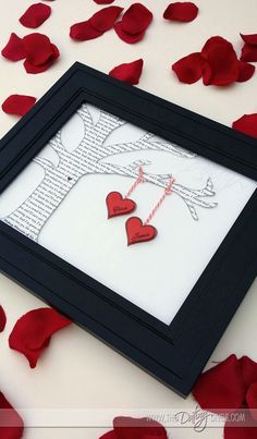 two red hearts hanging from a string in a black frame on a table with rose petals