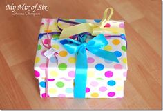 a gift wrapped in colorful polka dot paper with a blue ribbon and bow sitting on a wooden floor