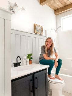 a woman sitting on top of a toilet next to a sink and bathtub in a bathroom