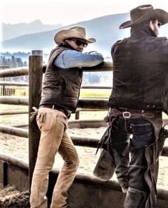 two men in cowboy hats standing next to each other near a fence with mountains in the background