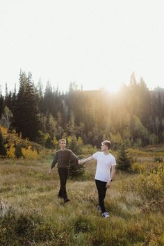 two people holding hands in the middle of a field with trees and mountains in the background