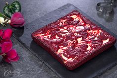 a square piece of red cake sitting on top of a black plate next to flowers