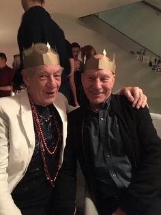 two older men sitting next to each other with crowns on their heads and people in the background