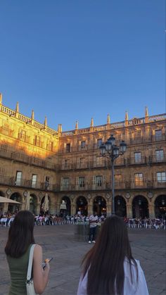 two women are looking at their cell phones in front of an old building with many people