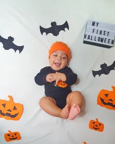 a baby wearing an orange hat and black bodysuit sitting in front of halloween decorations