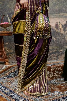 Make a statement with this luxurious velvet sari, elegantly paired with vertical color block panels in rich olive and purple hues. The design is accentuated by stunning mirror tassels along the border, adding a playful touch to the ensemble. Styled with an intricate mirror work blouse, this outfit beautifully blends traditional charm with contemporary flair, perfect for special occasions and celebrations. Note: Potli is for styling purposes only Velvet Sari, Mirror Tassels, Intricate Mirror, Mirror Work Blouse, Beach Wedding Guests, Jacket Cape, Indian Ethnic Wear, Mirror Work, Purple Hues