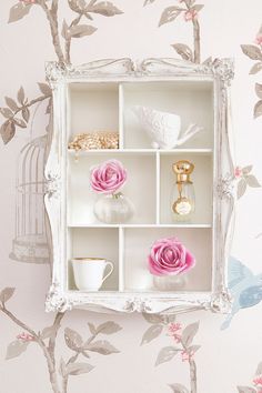 a white shelf with pink flowers and other items on it in front of a floral wallpaper