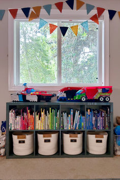 there is a book shelf with many books and toys in front of a large window