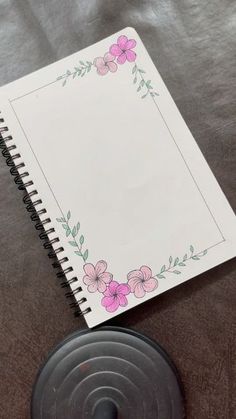 an open spiral notebook with pink flowers on it next to a black disk and pen