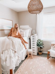 a woman sitting on top of a bed next to a white dresser and chair in a bedroom