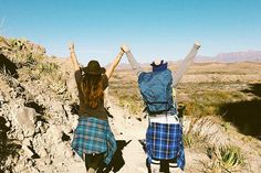 two women walking up a dirt path with their arms in the air and backpacks on their back