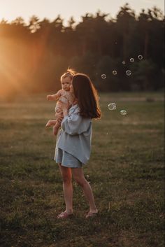 a woman holding a baby while standing in a field with soap bubbles flying around her