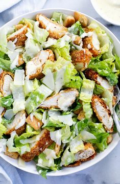 a white bowl filled with lettuce and chicken next to some dressing on a table