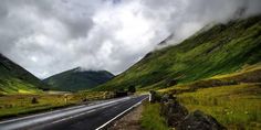 an empty road in the middle of mountains with clouds above it and grass on both sides