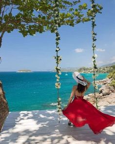 a woman in a red dress is sitting on a swing by the ocean and looking out at the water