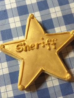 a cookie shaped like a star with the word sheriff spelled in gold icing on a blue and white checkered tablecloth