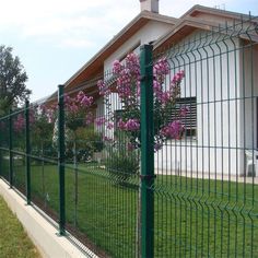 a green fence with pink flowers growing on it next to a white house and trees