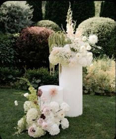 two tall white vases sitting on top of a green grass covered field next to bushes