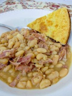 a white plate topped with beans and ham next to a piece of bread on top of a table