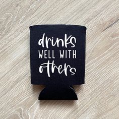 a black can holder with drinks well with others written on the front and bottom, sitting on a wooden surface