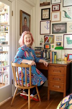 a woman with pink hair sitting at a desk in front of a wall full of pictures