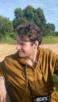 a man sitting in the grass with a camera around his neck and wearing a brown hoodie